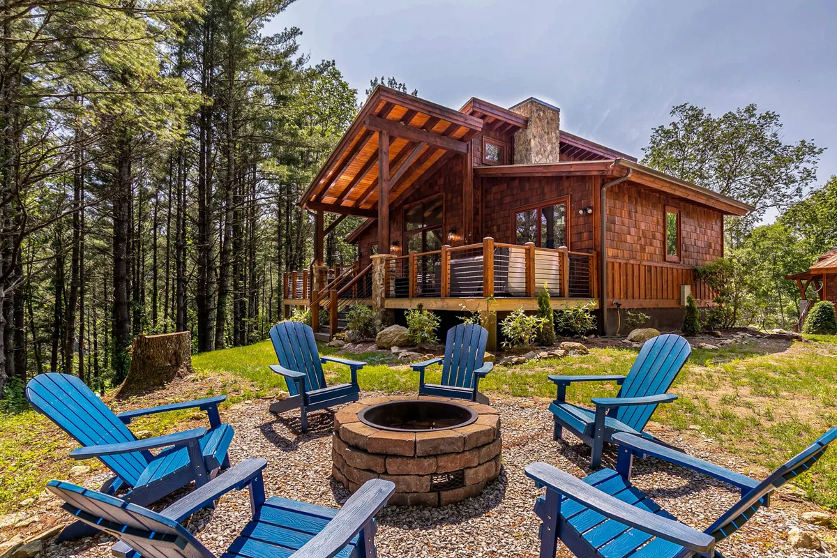 The image showcases the exterior of a charming wooden cabin nestled among tall pine trees atop a hill. The inviting fire pit, surrounded by vibrant blue Adirondack chairs, sits in the foreground, framed by the tranquil natural setting. The cabin's deck is visible, promising sunset views and peaceful mornings immersed in nature.