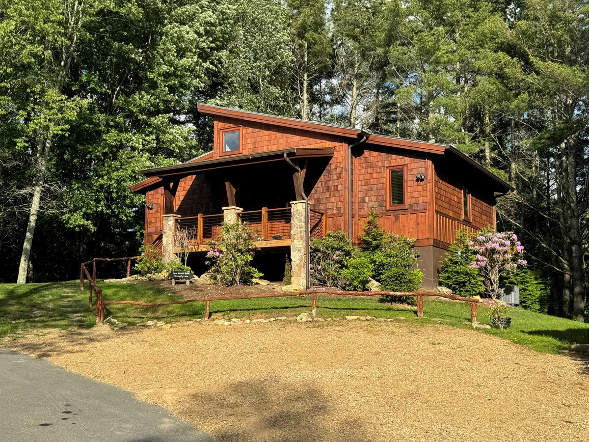 This image showcases a charming wooden cabin with prominent stone pillars, a landscaped garden, and a welcoming porch area. The scene exudes warmth and invites viewers to imagine relaxing outdoors, whether for morning coffee or evening gatherings. The lush greenery and tidy landscaping connect the cabin to its natural mountain surroundings.