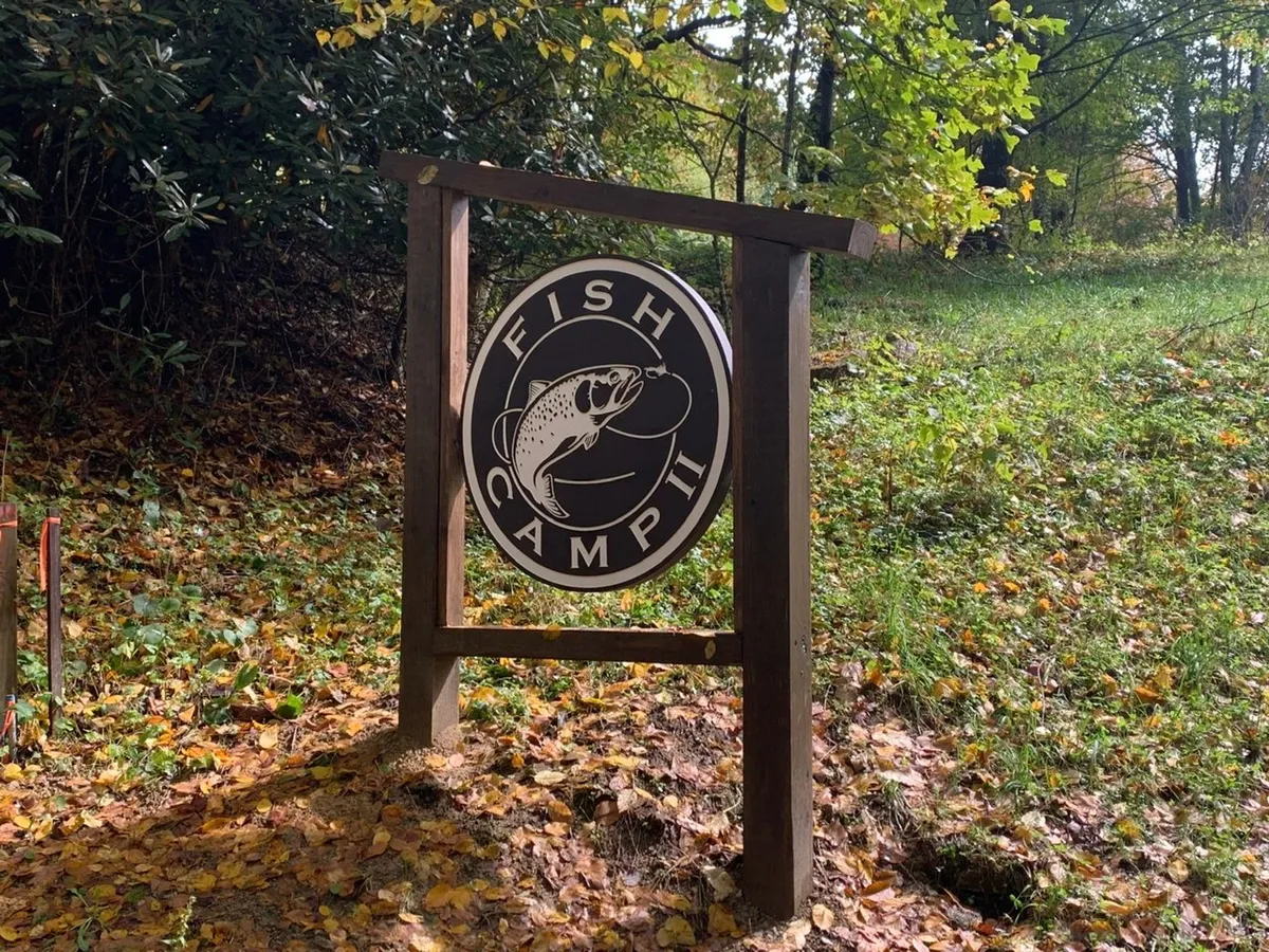 This image captures the Fish Camp entrance sign surrounded by lush foliage and colorful autumn leaves. The scene suggests an inviting, natural setting perfect for outdoor recreation like fishing, camping, hiking, and nature walks. The sign acts as a gateway to adventure within the woodland environment.