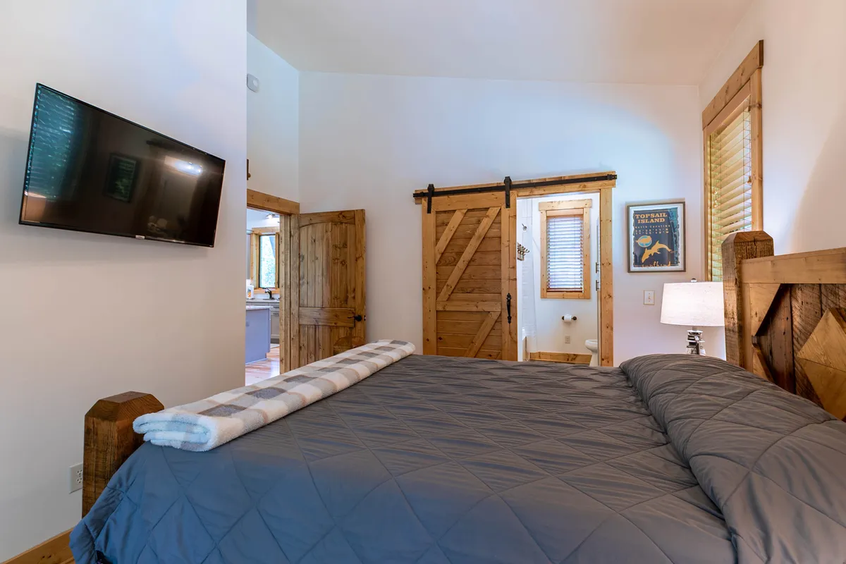 A serene bedroom featuring a comfortable bed with a gray quilt, a wooden sliding barn door, a wall-mounted television, and decorative artwork. Soft lighting from bedside lamps creates a restful, inviting atmosphere.