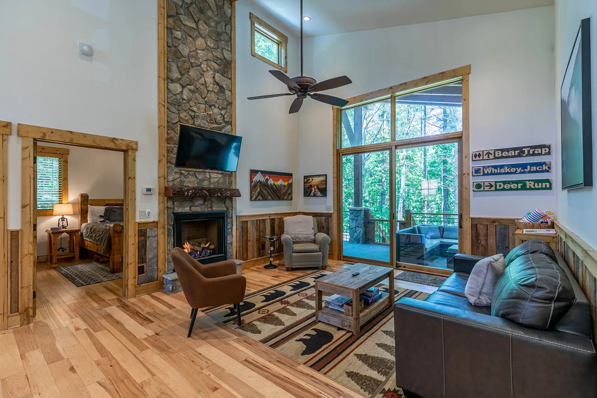 This image captures a spacious, open living area featuring a striking stone fireplace, large windows that bring in natural light, rustic wooden furniture, decorative rugs, and artwork on the walls. The scene suggests a warm atmosphere ideal for relaxation and social interaction.