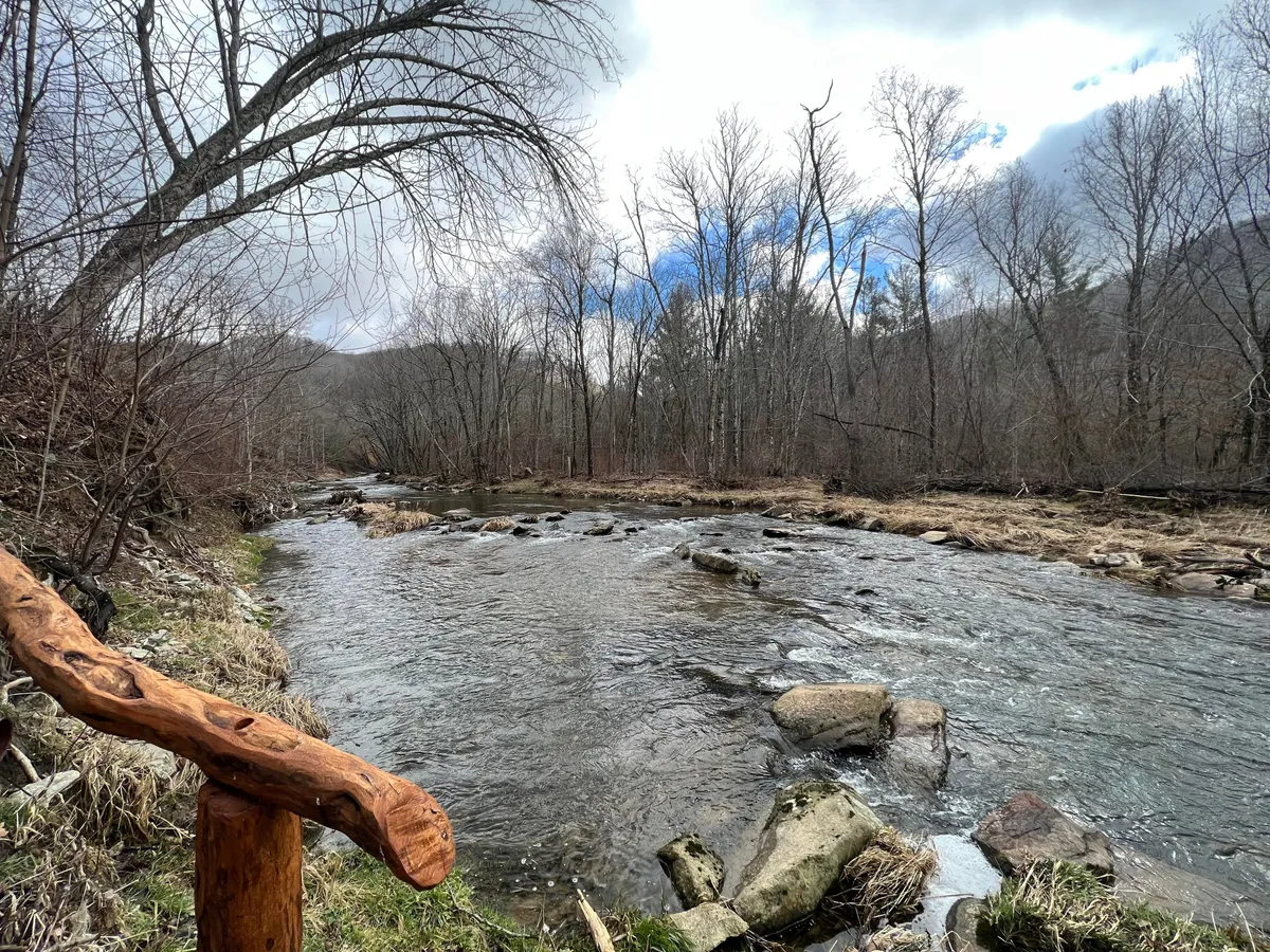A peaceful scene of a flowing river with a wooden railing and rocky riverbank, bordered by bare trees. The area is ideal for nature walks, fishing, and quiet family moments, especially during sunset with reflections on the water.