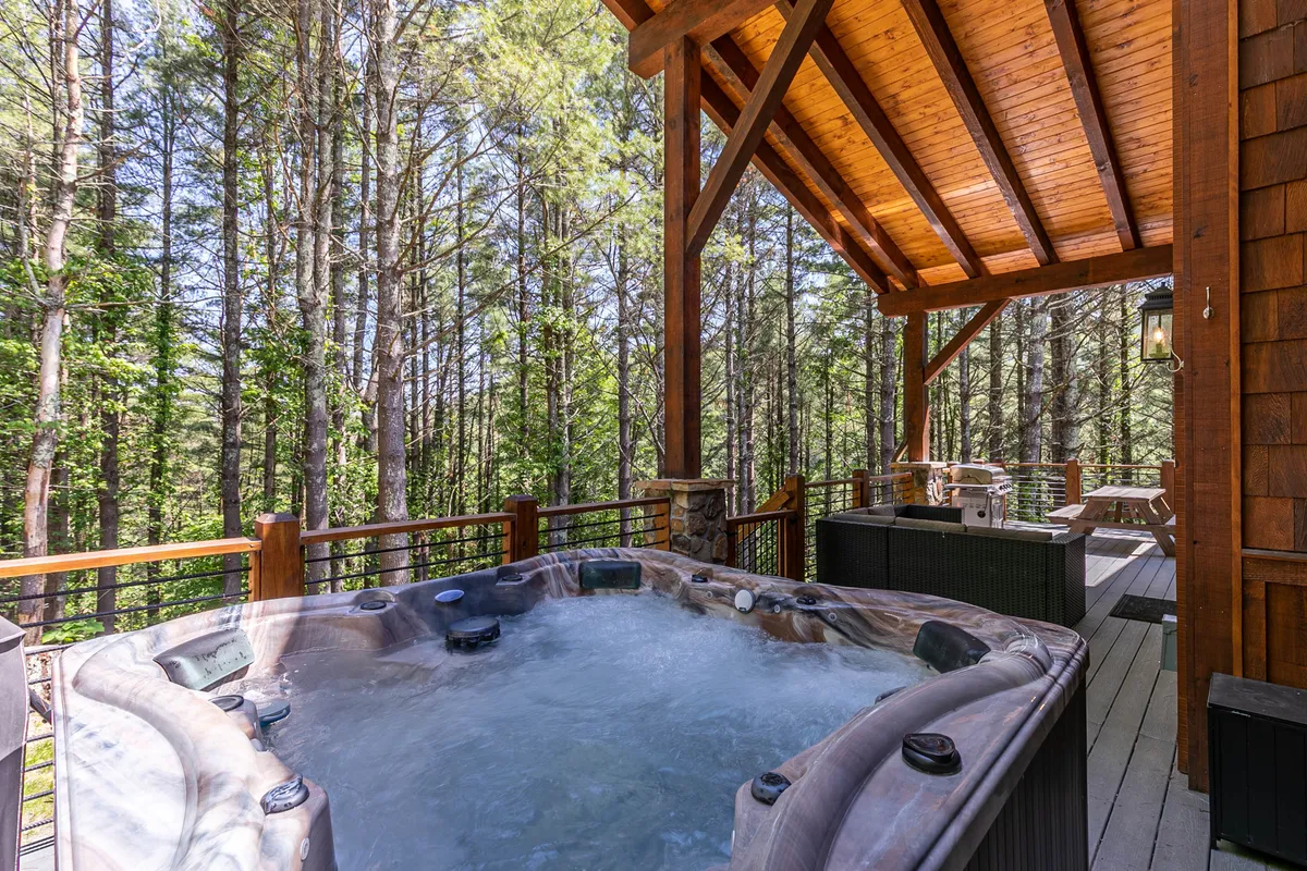 A luxurious hot tub set on a wooden deck, surrounded by tall trees that provide privacy and a sense of immersion in nature. The deck is spacious and designed for both intimate relaxation and social gatherings, reinforcing the feeling of year-round enjoyment in a secluded setting.