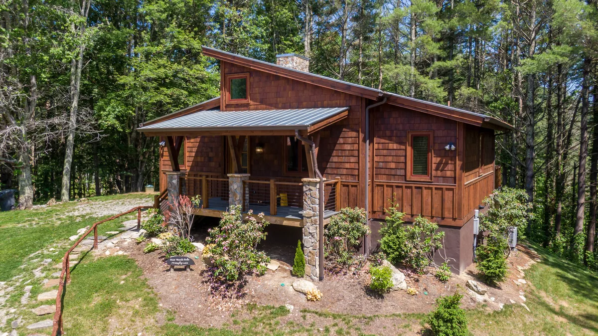 The image captures a peaceful wooden porch with sturdy stone pillars, surrounded by lush greenery and decorative flowers. The scene suggests a tranquil setting for sipping coffee at sunrise or engaging in intimate evening conversations under the stars.